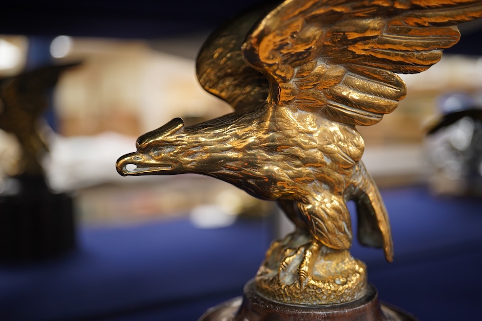 Two brass car mascots in the form of eagles, one mounted on a threaded radiator cap, the other mounted on a wooden base, the larger with a wingspan of 31cm. Condition - fair, one eagle with bent wing.